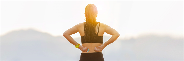 Woman holding back wearing athletic wear, blurred background of hills and sun